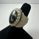 Handwoven ring with a large polished black and white stone center, surrounded by beige braided fibers with a thin turquoise accent band.