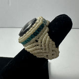 Handwoven ring featuring cream-colored macrame with teal accents and a large, faceted dark stone centerpiece, displayed on a black velvet finger stand.