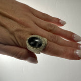 Handmade woven ring with beige thread encasing a polished black stone with white specks, worn on a hand with light pink glossy nails.