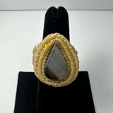 Teardrop-shaped gray banded agate stone set in a cream-colored woven ring band on a black finger display stand