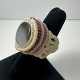 Handwoven ring featuring a smooth, oval gray stone centerpiece surrounded by cream and mauve woven bands, displayed on a black ring stand.