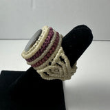 Handmade woven ring featuring cream and maroon intertwined threads, topped with a smooth round stone, displayed on a black ring stand.
