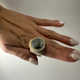 Hand wearing a handmade woven ring with a central smooth gray stone and beige crocheted or woven band, shown against a plain light background.