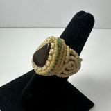 Handmade beige woven ring with a teardrop-shaped dark stone in the center, displayed on a black velvet ring holder against a light background.