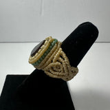 Handwoven beige cord ring featuring a dark oval stone centerpiece and subtle green accents, displayed on a black velvet ring holder against a plain background.