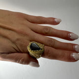 Hand with neutral-toned manicured nails wearing a beige woven ring featuring a large, polished black triangular stone on the middle finger.