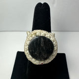 Cream-colored woven ring band featuring a round dark stone with subtle grayish texture, displayed on a black vertical ring stand.