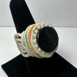 Handwoven cream-colored ring with delicate orange and teal thread accents, featuring a smooth, round black stone centerpiece, displayed on a black velvet stand.