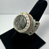 Handmade woven ring with a textured gray stone center and an off-white braided band accented by subtle red and blue threads, displayed on a black stand.