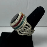 Woven ring featuring a dark stone centerpiece surrounded by coils of white cord interspersed with orange, red, and teal woven bands on a black display stand.