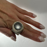 Hand wearing a macrame woven ring featuring a round black and white stone, shown against a plain white background with glossy pale pink and white nails.