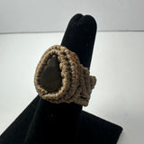 Handmade woven ring featuring beige cord tightly wrapped around a teardrop-shaped dark stone, displayed on a black velvet finger stand.