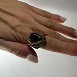 Hand wearing a handmade woven ring featuring a dark polished stone set in thick beige cord wrapping on the ring finger against a white background.