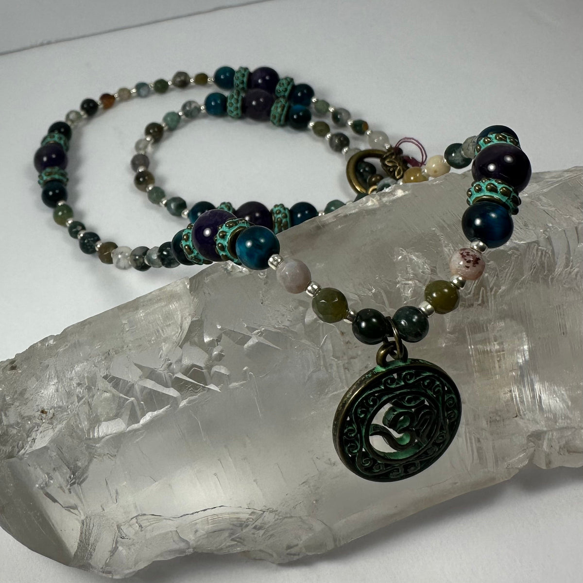 Bronze patina circular pendant with 'Om' symbol on a multi-colored beaded necklace resting on a translucent quartz crystal piece.