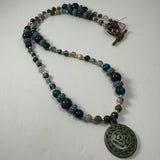 Mixed bead necklace featuring round beads in teal, purple, and earthy tones with a bronze patina circular pendant embossed with the 'Om' symbol and a toggle clasp.
