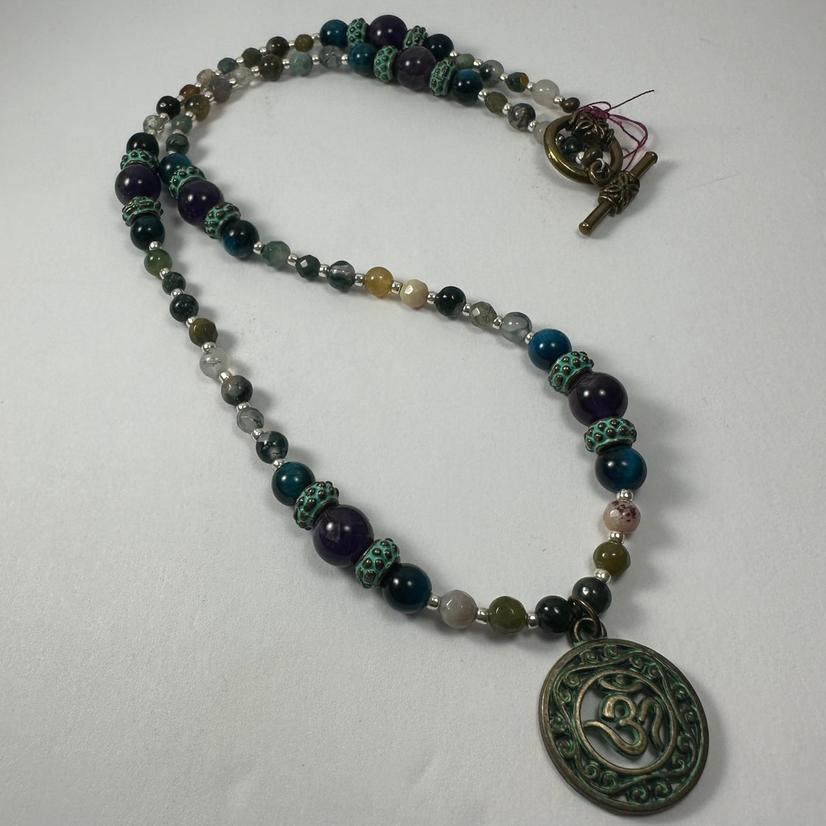 Mixed bead necklace featuring round beads in teal, purple, and earthy tones with a bronze patina circular pendant embossed with the 'Om' symbol and a toggle clasp.