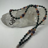 Beaded necklace with dark green, amber, and purple beads displayed on a clear quartz crystal, featuring a silver 'Om' pendant near one end.