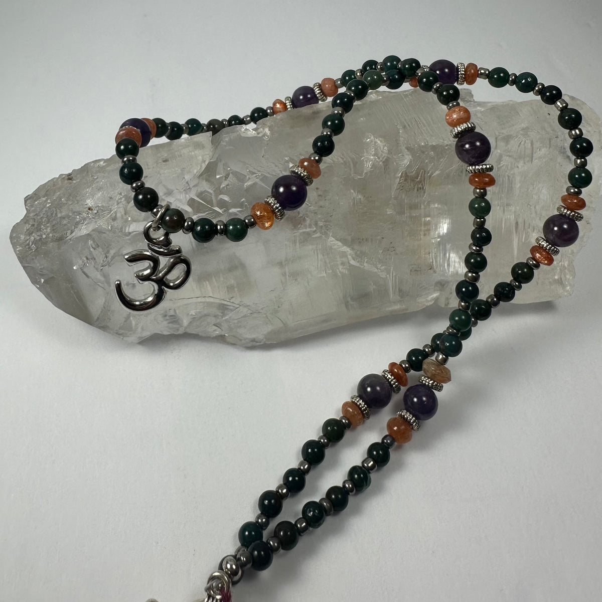Beaded necklace with dark green, amber, and purple beads displayed on a clear quartz crystal, featuring a silver 'Om' pendant near one end.