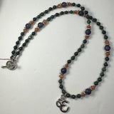 Beaded necklace with dark green and purple beads accented by orange spacers, featuring a silver 'Om' pendant and a silver toggle clasp, displayed on a white background.