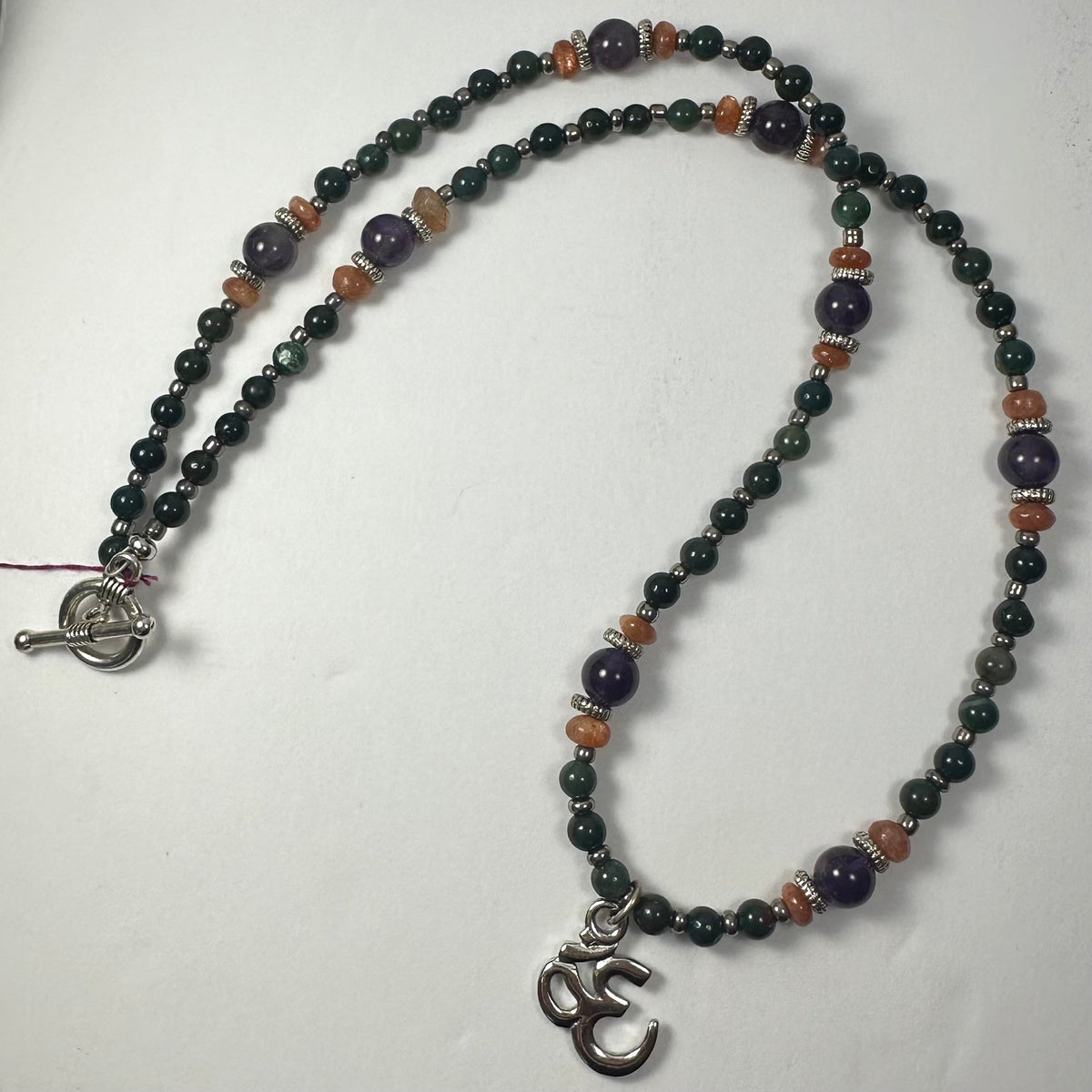 Beaded necklace with dark green and purple beads accented by orange spacers, featuring a silver 'Om' pendant and a silver toggle clasp, displayed on a white background.