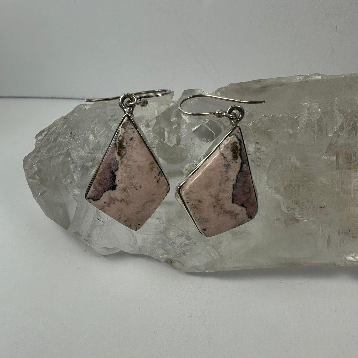 Pair of rhodonite gemstone earrings with pink and black veining set in silver, displayed on a clear quartz crystal cluster against a white background.