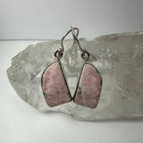 Pair of drop earrings featuring polished pink rhodonite gemstones with silver metal hooks, displayed resting on a clear quartz crystal cluster.