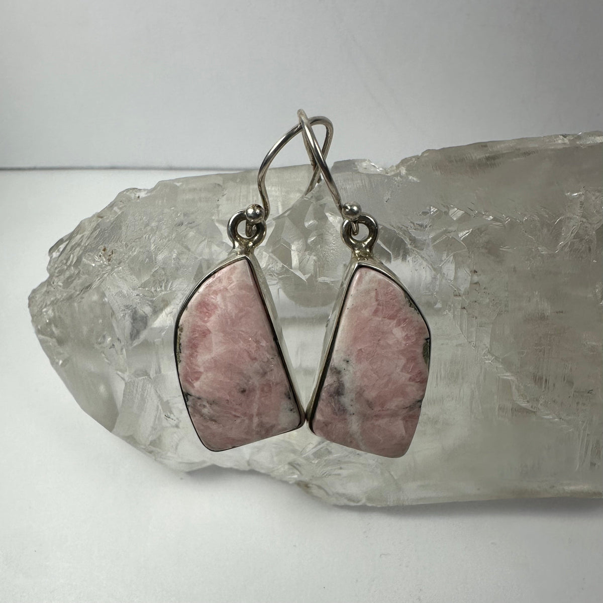 Pair of drop earrings featuring polished pink rhodonite gemstones with silver metal hooks, displayed resting on a clear quartz crystal cluster.
