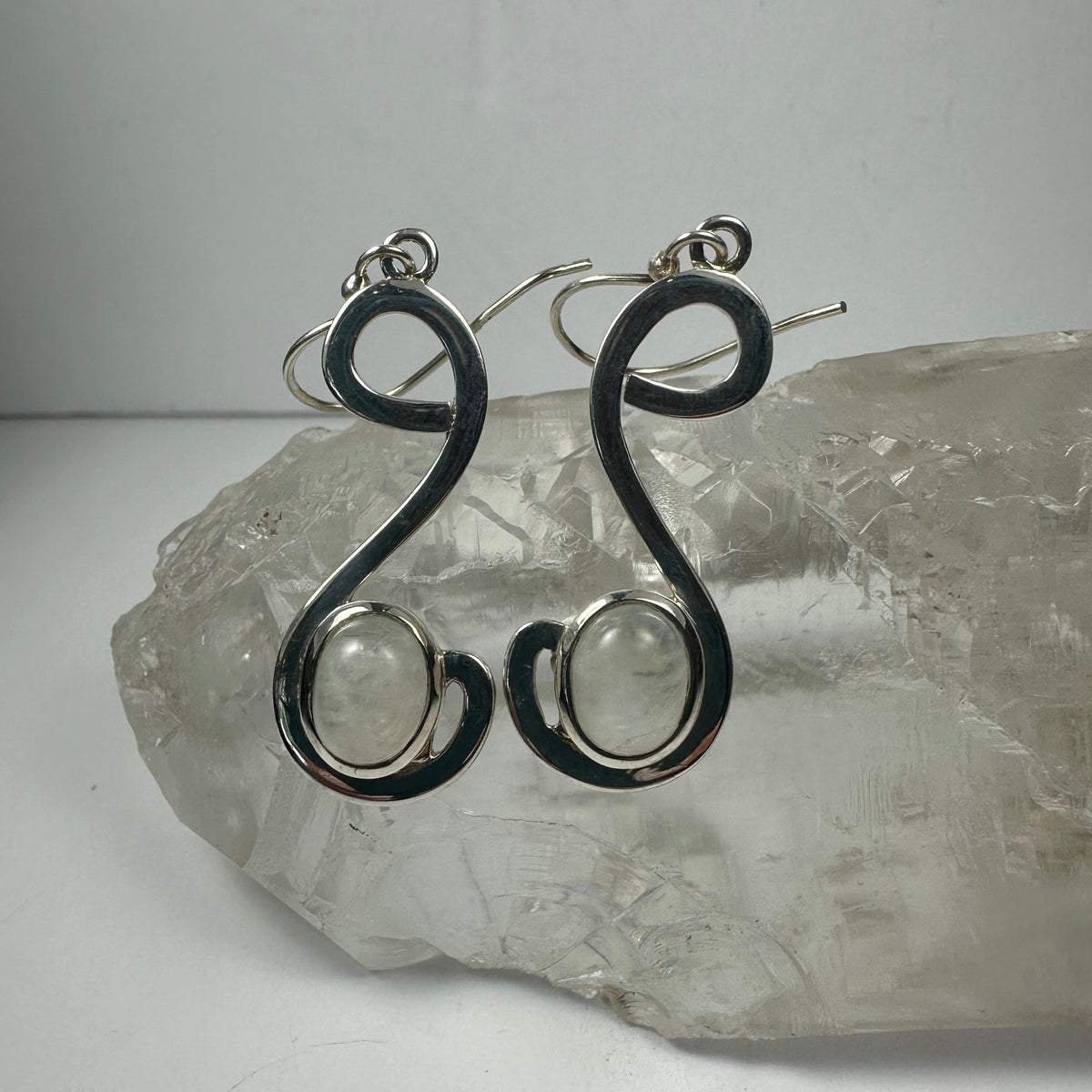 Pair of silver earrings with an elegant, curved 'S' shape design, each featuring a round moonstone set near the lower curve, displayed on a clear quartz crystal.