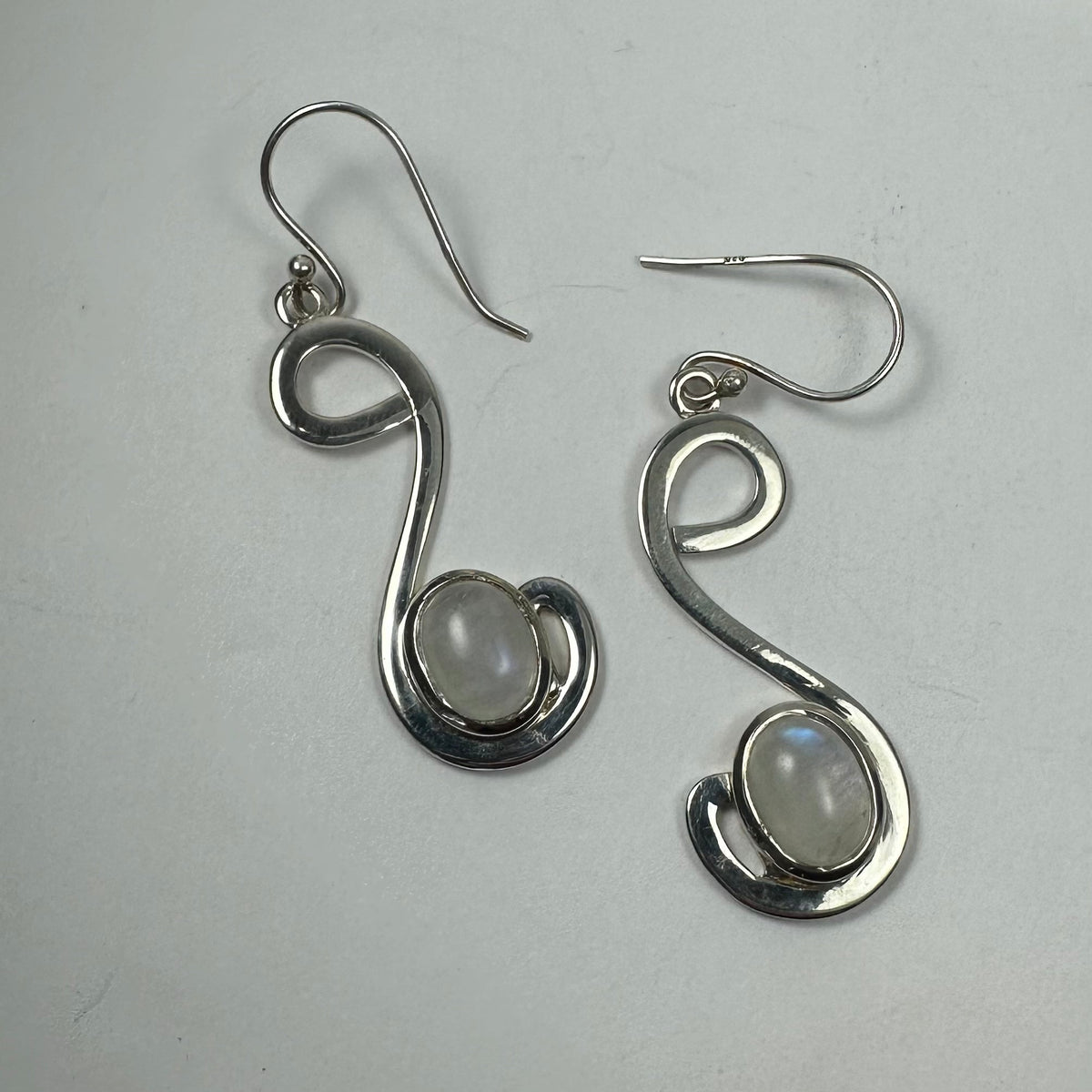 Pair of silver earrings with a freeform looping design, each featuring a smooth, oval moonstone set near the curve's base on a light gray background.