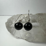 Pair of black onyx drop earrings with polished round stones set in silver hooks, displayed on a large translucent crystal with textured surface.