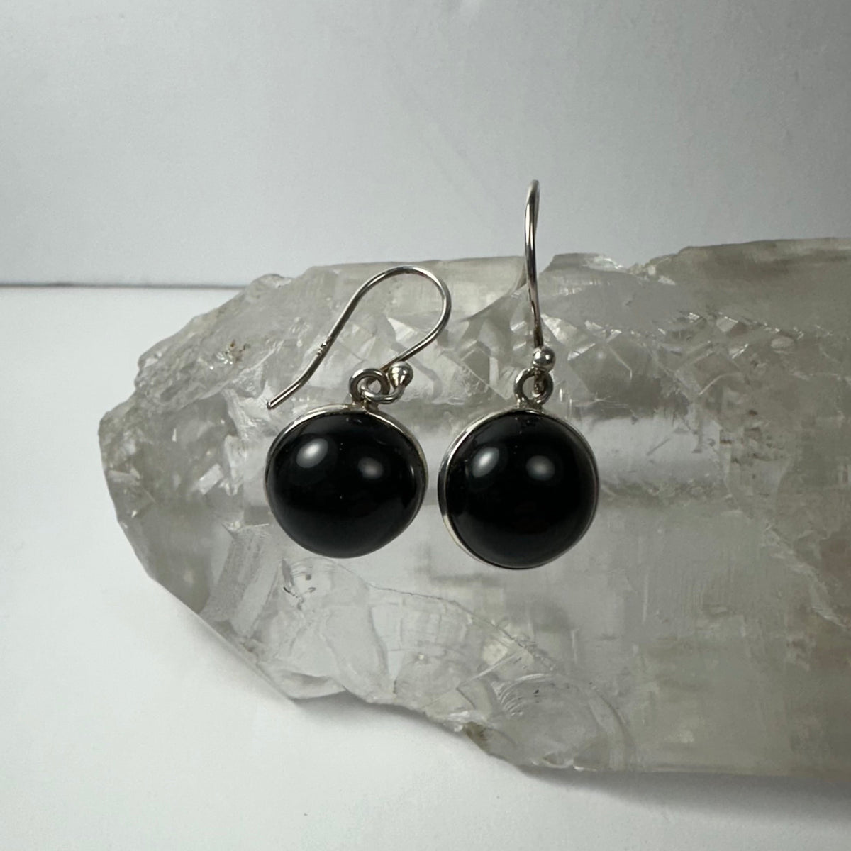 Pair of black onyx drop earrings with polished round stones set in silver hooks, displayed on a large translucent crystal with textured surface.