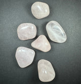 Six large, polished tumbled stones in pale pink and translucent white hues arranged loosely on a dark gray background.