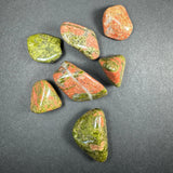 Seven polished tumbled stones in green and reddish-orange hues with smooth, rounded shapes on a dark gray background.