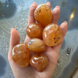 Hand holds six polished orange-brown tumbled stones with smooth, glossy surfaces and dark inclusions, shown against a textured blue-gray background with circular patterns.