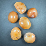 Six polished tumbled stones in warm tones of orange, amber, and beige with smooth surfaces and subtle internal patterns, arranged on a gray background.
