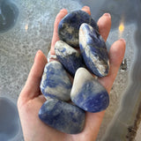 Hand holding six large smooth tumbled stones with blue and white marbled patterns, set against a background of a light gray agate surface.