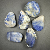 Six polished tumbled stones with blue, white, and gray marbled patterns arranged on a dark gray surface, each stone showing unique irregular shapes and color distribution.