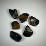 Six polished tumbled stones with smooth surfaces, predominantly black with brown and gold streaks, arranged loosely on a white background.