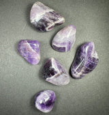 Six polished amethyst stones with shades of purple and white, featuring smooth surfaces and varied rounded triangular shapes, arranged on a gray background.