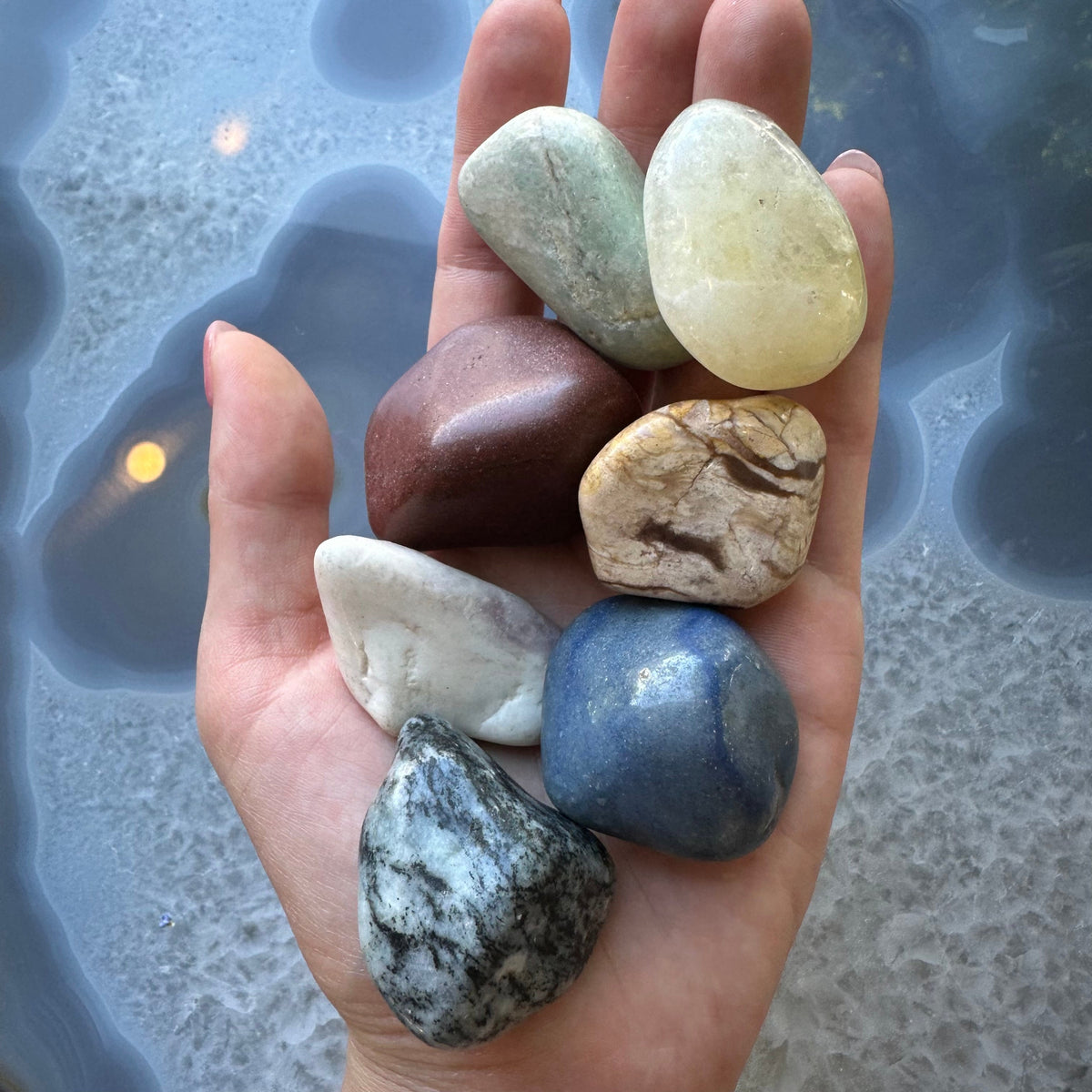 Hand holding seven large polished tumbled stones in various colors including green, yellow, red, brown, white, blue, and gray with black streaks, against a patterned blue-gray surface.