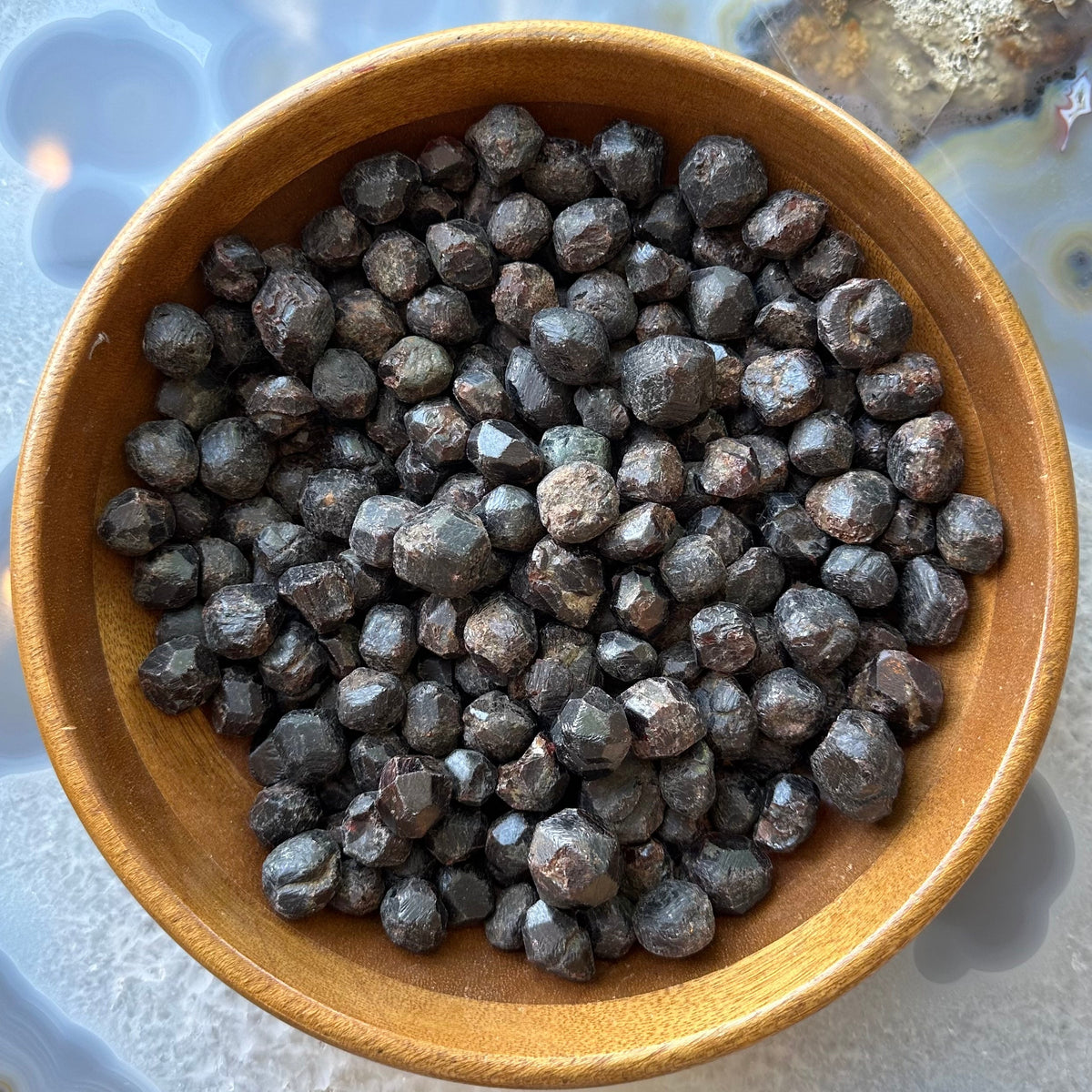 Wooden bowl filled with small, dark red-brown garnet stones with a rounded, slightly faceted surface, displayed on a blue and gray patterned stone slab.