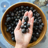 Hand holding multiple dark reddish-brown faceted garnet stones over a wooden bowl filled with similar stones on a polished blue-grey surface.