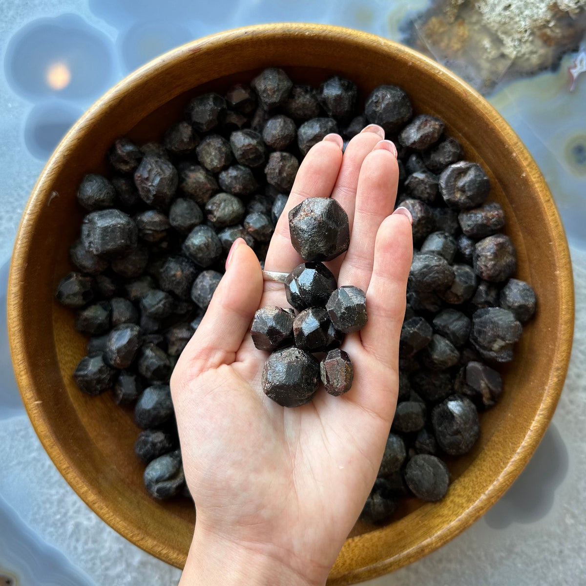 Hand holding multiple dark reddish-brown faceted garnet stones over a wooden bowl filled with similar stones on a polished blue-grey surface.