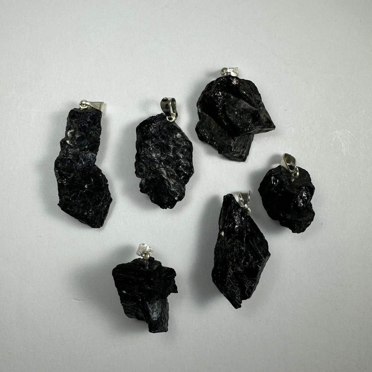 Six irregularly shaped black crystal pendants with silver bails arranged on a plain light gray background, highlighting their glossy and textured surfaces.