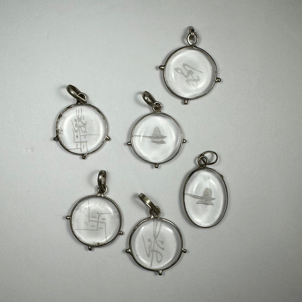 Six round and oval clear crystal pendants with various abstract and symbolic carvings, each set in simple metal frames with small loops for hanging.