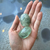 Hand holding two translucent light green fluorite tumbled stones with smooth, rounded shapes and natural internal fractures, against a textured blue-gray surface.