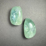 Two translucent tumbled rainbow fluorite stones with green and blue hues, placed on a gray textured background showing natural internal patterns.