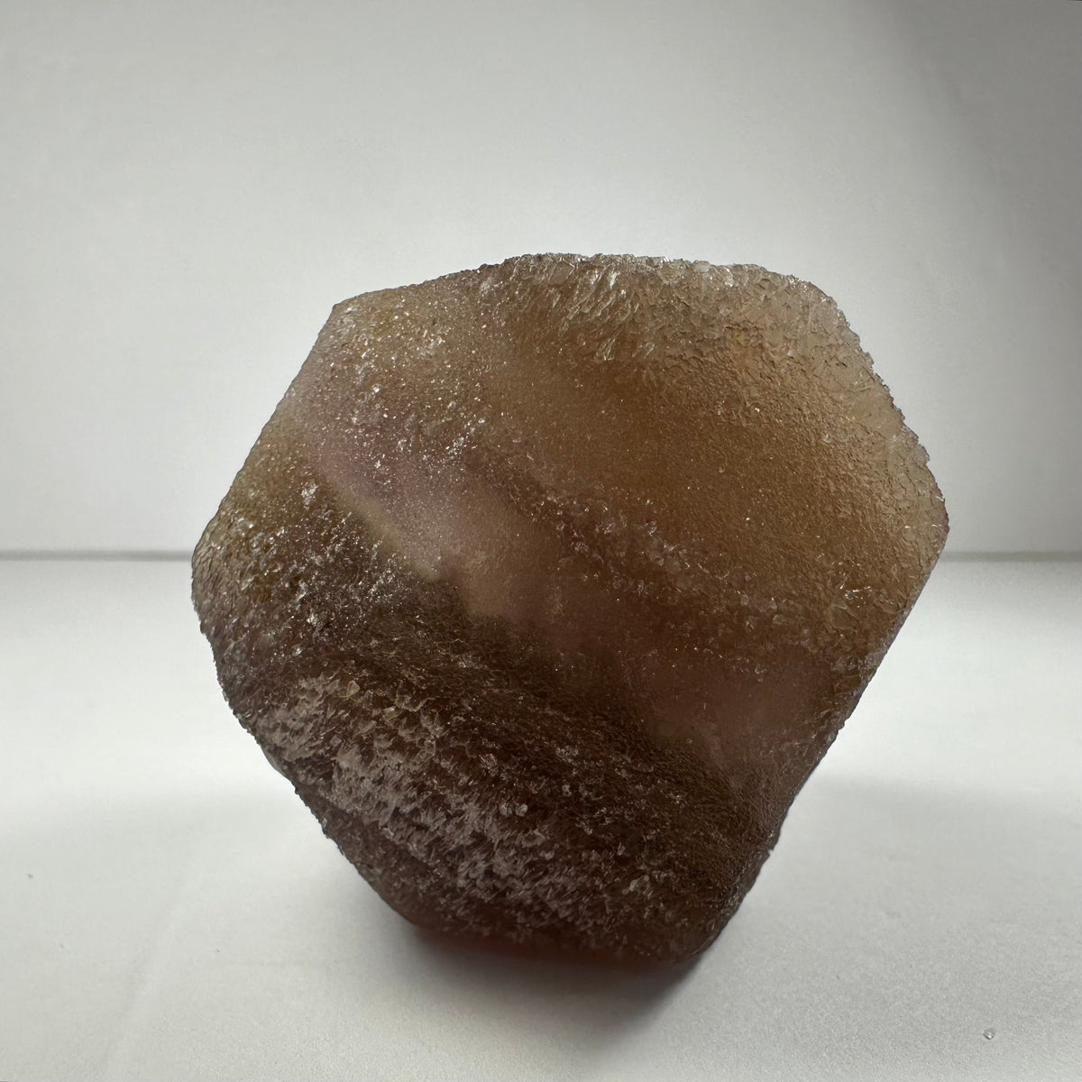 Close-up of a translucent brown fluorite dodecahedron with a textured, crystalline surface on a white background, showing natural irregularities and subtle light reflections.