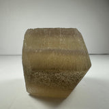 Translucent beige fluorite dodecahedron crystal with a matte texture and subtle striations, resting on a white surface against a plain white background.