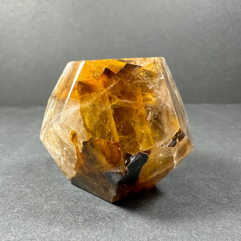 Yellow hematite quartz dodecahedron crystal carving with translucent honey and amber tones and dark inclusions, displayed against a gray backdrop.